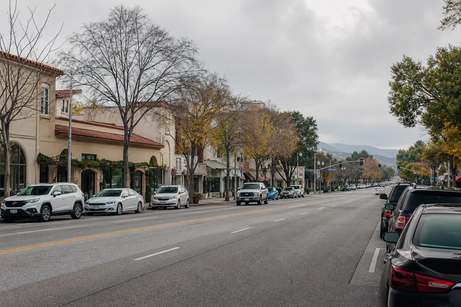Overcast main road through town with Spanish-style storefronts, bare and autumn trees, parked cars along both curbs and hills in the distance