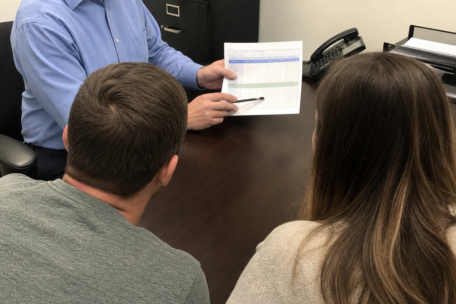 Agent in a blue dress shirt holds a printed comparison chart and pen at a dark wood desk while a couple seated across, seen from behind, looks on; filing cabinet and desk phone beh