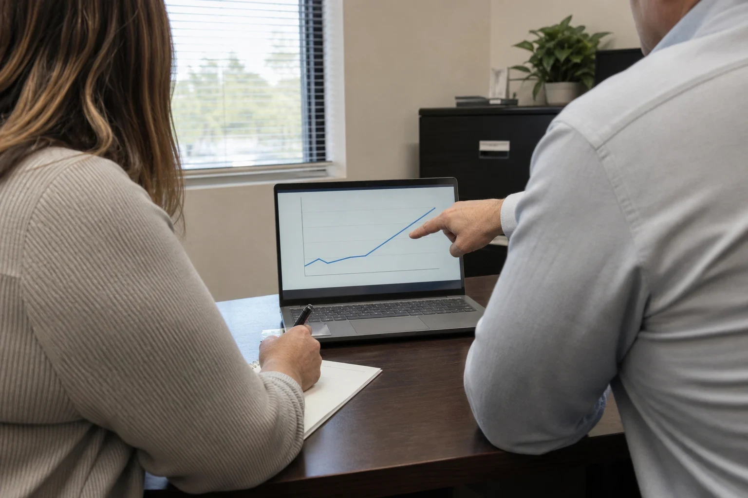 Over-the-shoulder view of an agent pointing to a simple line chart on a laptop while a client holds a pen over a notepad at an office desk near a window and black filing cabinet